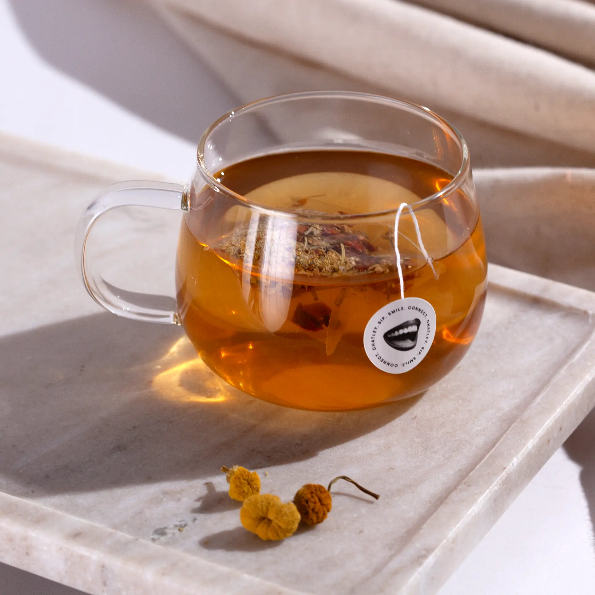 Chatley herbal tea steeping in a glass cup, its Chatley teabag tag resting on a marble tray with chamomile buds